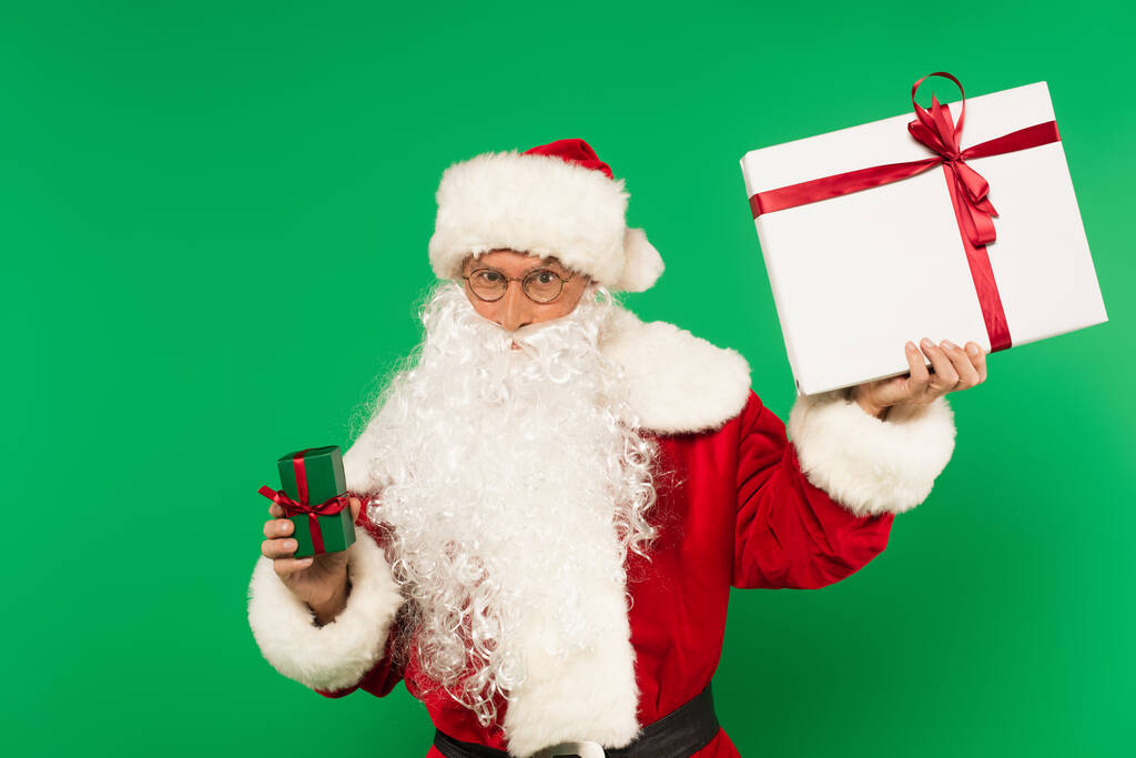 Kerstman houdt cadeautjes vast en kijkt naar camera geïsoleerd op groen - Foto, afbeelding