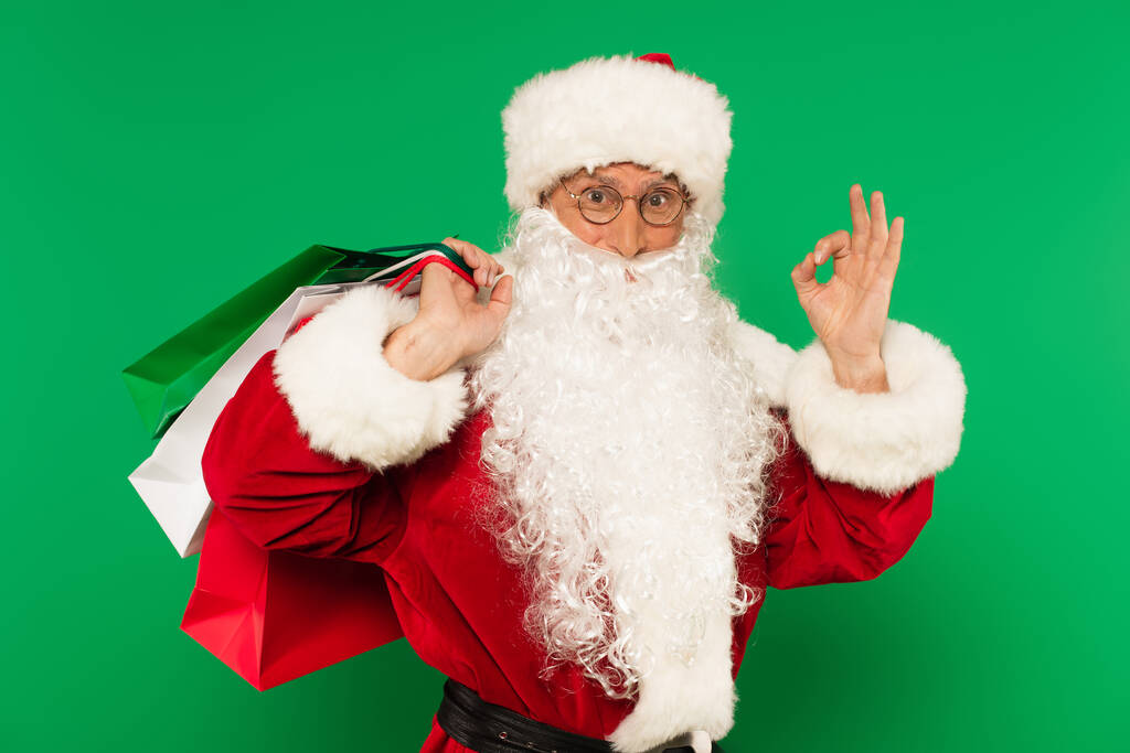 Santa Claus με τσάντες ψώνια δείχνει εντάξει χειρονομία απομονώνονται σε πράσινο - Φωτογραφία, εικόνα