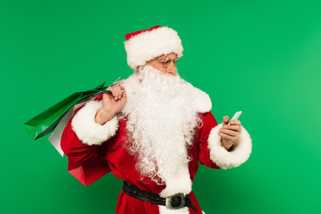 Santa claus with shopping bags using smartphone isolated on green - Photo, Image