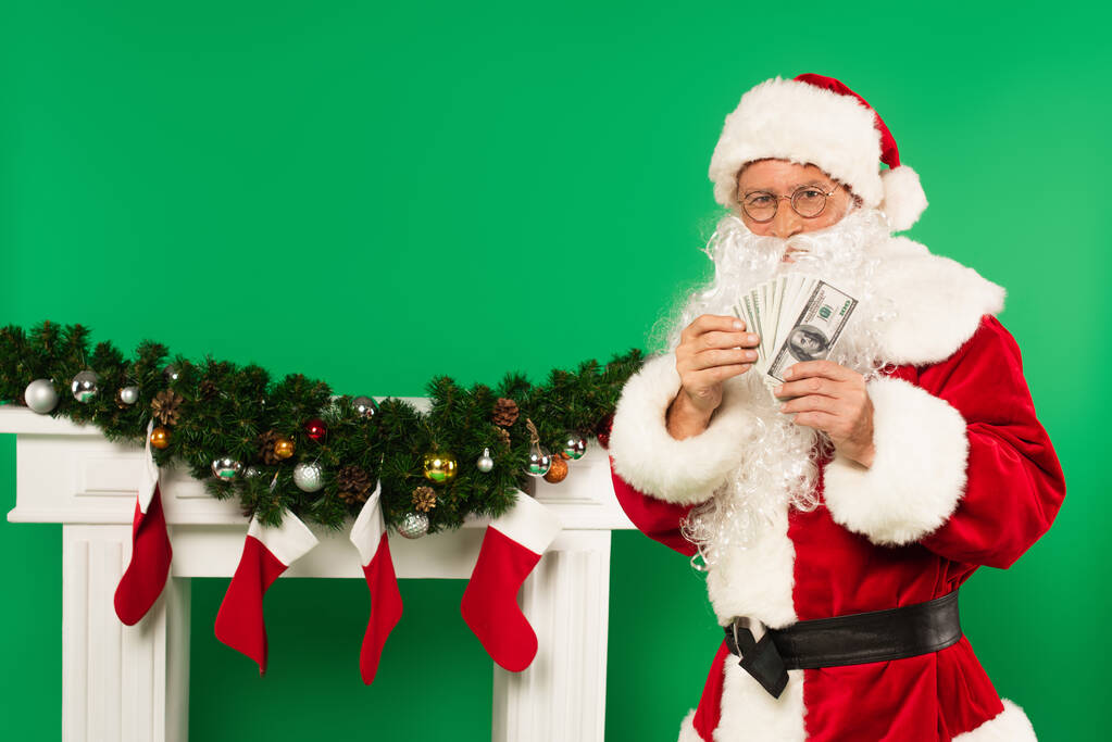 Santa Claus držení peněz v blízkosti zdobené krb s vánoční punčochy na zeleném pozadí - Fotografie, Obrázek
