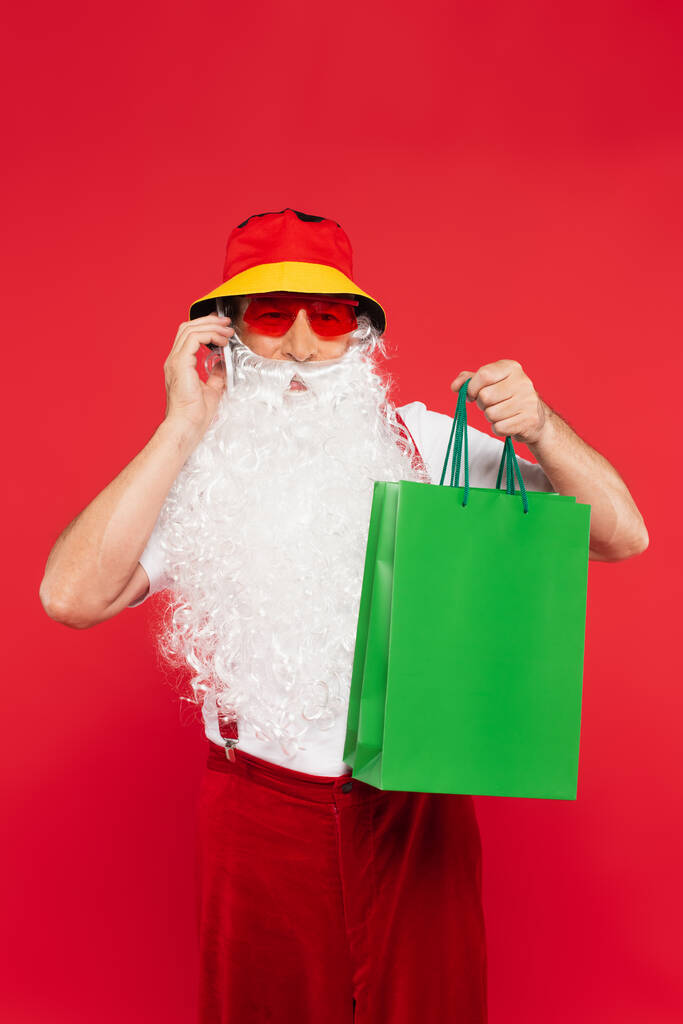 Santa Claus v Panamě a sluneční brýle mluví na smartphone a drží nákupní tašky izolované na červené  - Fotografie, Obrázek