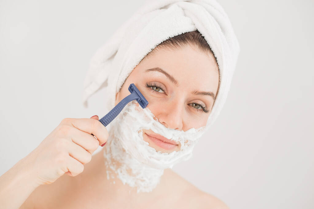Cheerful caucasian woman with a towel on her head and shaving foam on her face holds a razor on a white background - Photo, Image