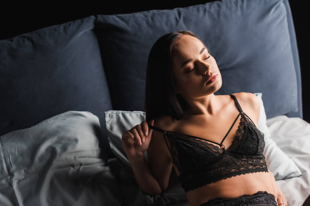 Αισθησιακή γυναίκα με δαντελωτά εσώρουχα ξαπλωμένη στο κρεβάτι σε μαύρο φόντο - Φωτογραφία, εικόνα