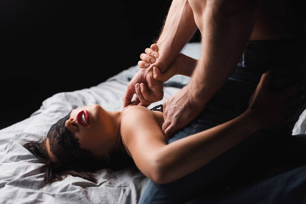 man aanraken gepassioneerde vriendin in kant masker op bed geïsoleerd op zwart - Foto, afbeelding