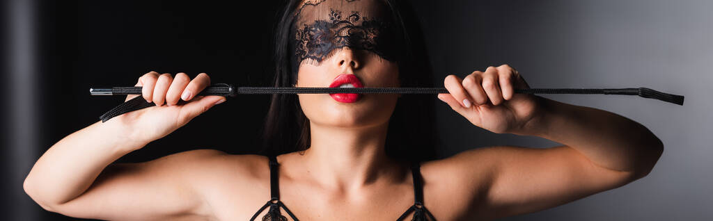 Sensuele vrouw in kant masker en beha houden spanking paddle op zwarte achtergrond, banner  - Foto, afbeelding