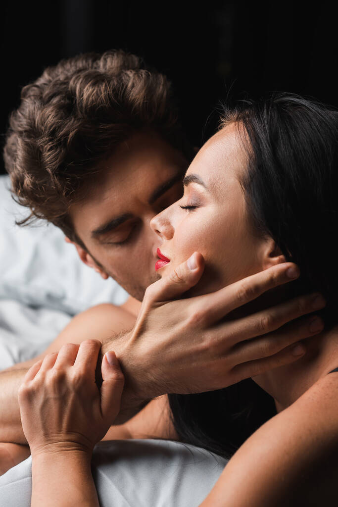 mężczyzna całowanie i dotykanie sexy brunetka dziewczyna na łóżko izolowane na czarny - Zdjęcie, obraz