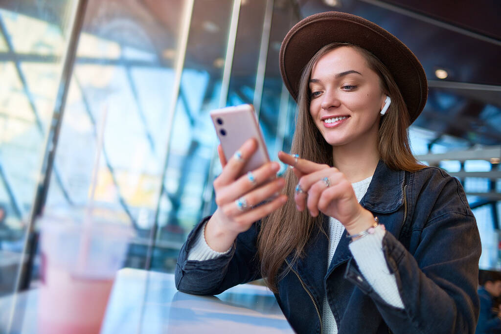 Portré stílusos trendi boldog hipster alkalmi nő kalap vezeték nélküli fülhallgatóval és mobiltelefon egy kávézóban. Modern emberek digitális életmód - Fotó, kép