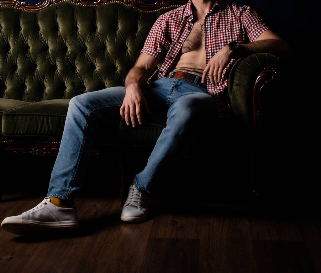 Egy felismerhetetlen férfi kigombolt ingben ül egy kanapén egy sötét szobában.. - Fotó, kép