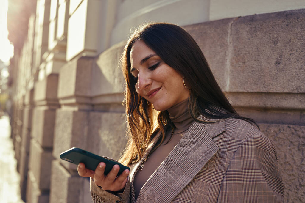 Hyvä nainen seisoo rakennuksen vieressä hymyillen katsoessaan älypuhelintaan - Valokuva, kuva