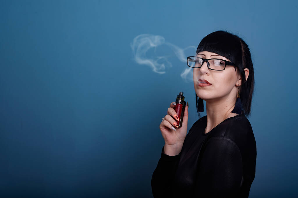 A beautiful girl with dark hair and glasses exquisitely smokes an electronic cigarette on a dark background - Photo, Image