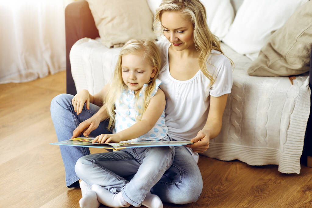 Счастливая семья. Молодая блондинка читает книгу своей милой дочери, сидя на деревянном полу в солнечной комнате. Концепция материнства - Фото, изображение