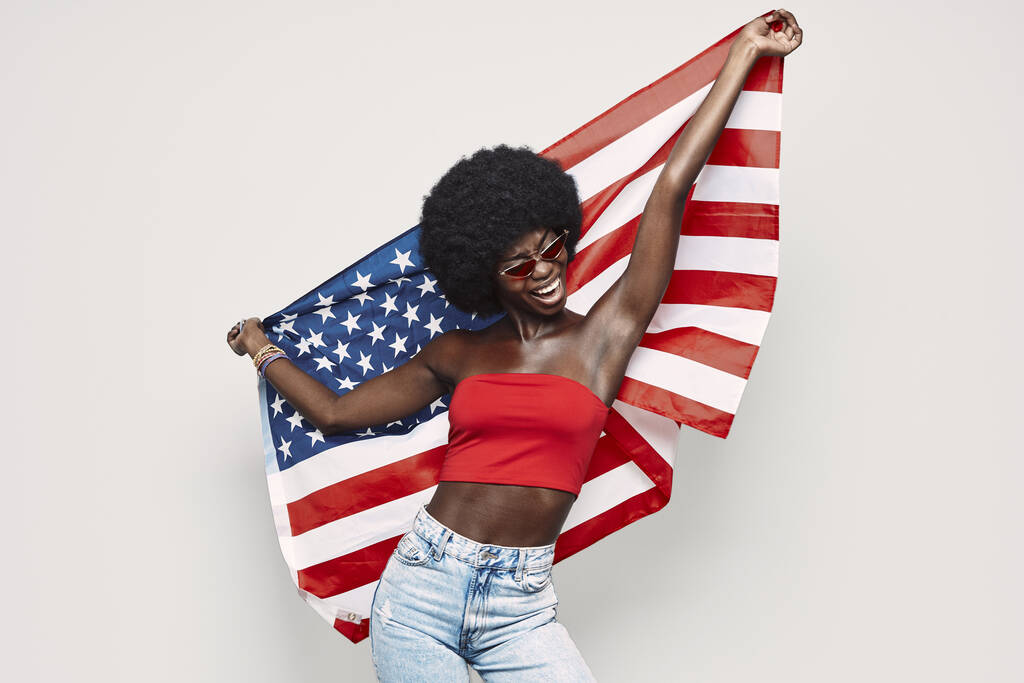 Щаслива молода африканка, що несе американський прапор і посміхається, стоячи проти сірого фону. - Фото, зображення