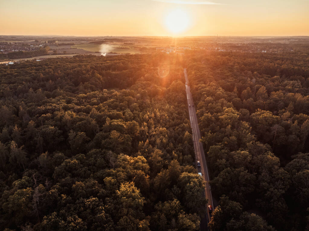 Uitzicht vanaf een hoogte naar beneden naar de grond op het bos en de weg in het bos. De zonsondergang en de stad zijn op de achtergrond te zien. - Foto, afbeelding