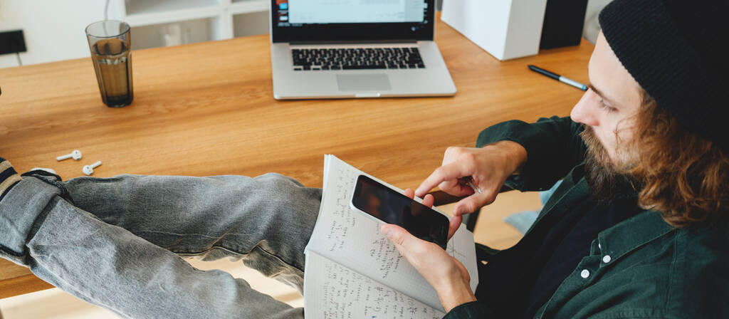 Szakállas főiskolai hallgató készül át vizsga otthon okostelefon. A fiatalember jegyzetel, miközben online tanul. Széles kép - Fotó, kép