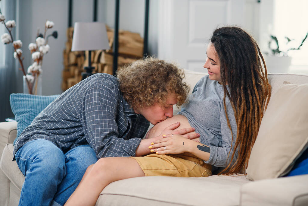 Муж трогает беременный животик своей очаровательной радостной жены, которая лежит на диване в уютной гостиной - Фото, изображение