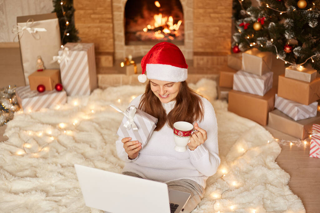Horní pohled na mladou dospělou ženu sedící na podlaze se svým laptopem, s veselým výrazem, pijící čaj z hrnku při videohovoru, ukazující dárkovou krabici na webovou kameru. - Fotografie, Obrázek