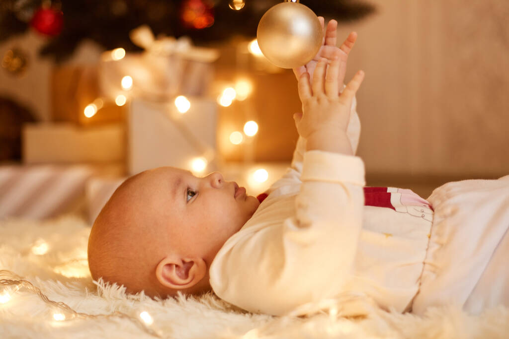 Side view portrait of cute charming infant girl lying on floor on soft carpet playing with ball from Christmas tree, new year eve celebration, indoor shot. - Photo, Image