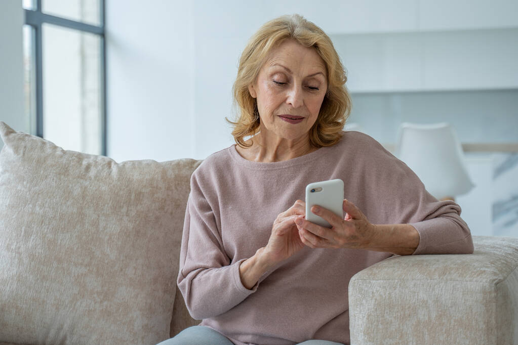 Une charmante grand-mère assise sur le canapé dans le salon de la maison, faisant des achats en ligne avec un smartphone dans ses mains - Photo, image