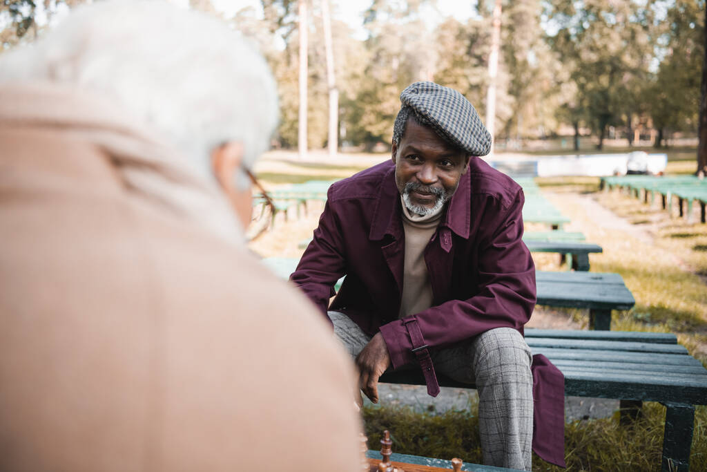 Uomo anziano afroamericano che guarda un amico offuscato vicino agli scacchi nel parco  - Foto, immagini