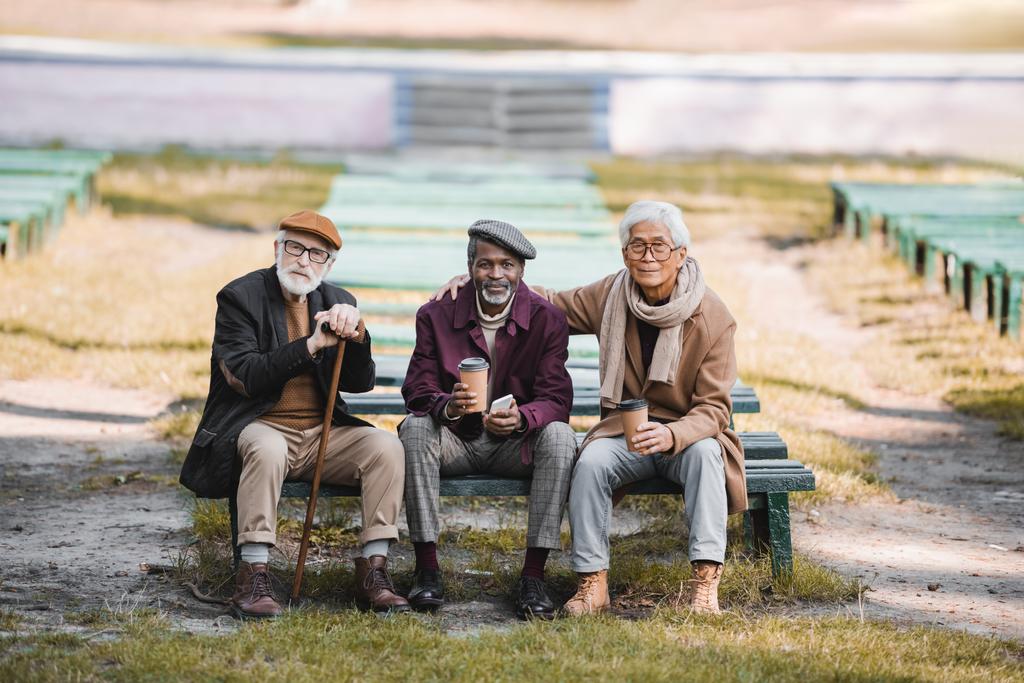 Wieloetniczni starsi mężczyźni z papierowymi kubkami i smartfonem siedzący na ławce obok przyjaciela z chodzącą laską  - Zdjęcie, obraz