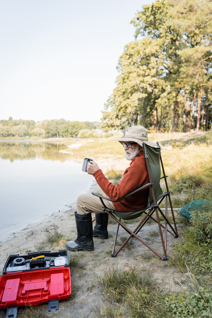 Vanhempi mies kalastusasussa pitämässä lämpökuppia työkalupakin ja järven lähellä  - Valokuva, kuva