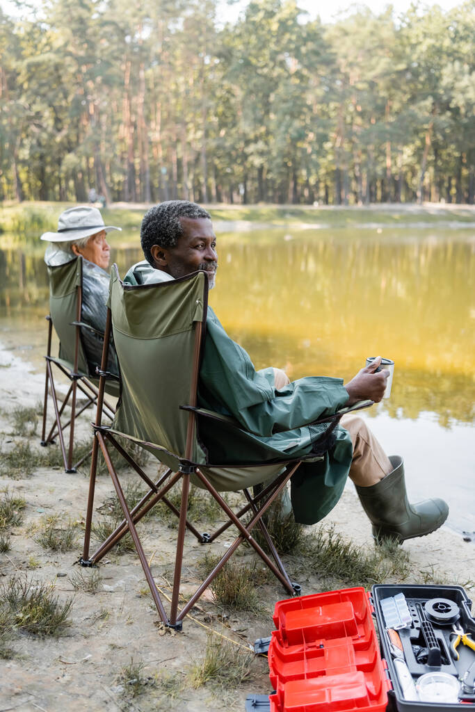 アフリカ系アメリカ人の男を保持熱カップ近くアジアの友人で釣り服で近くツールボックスと湖  - 写真・画像