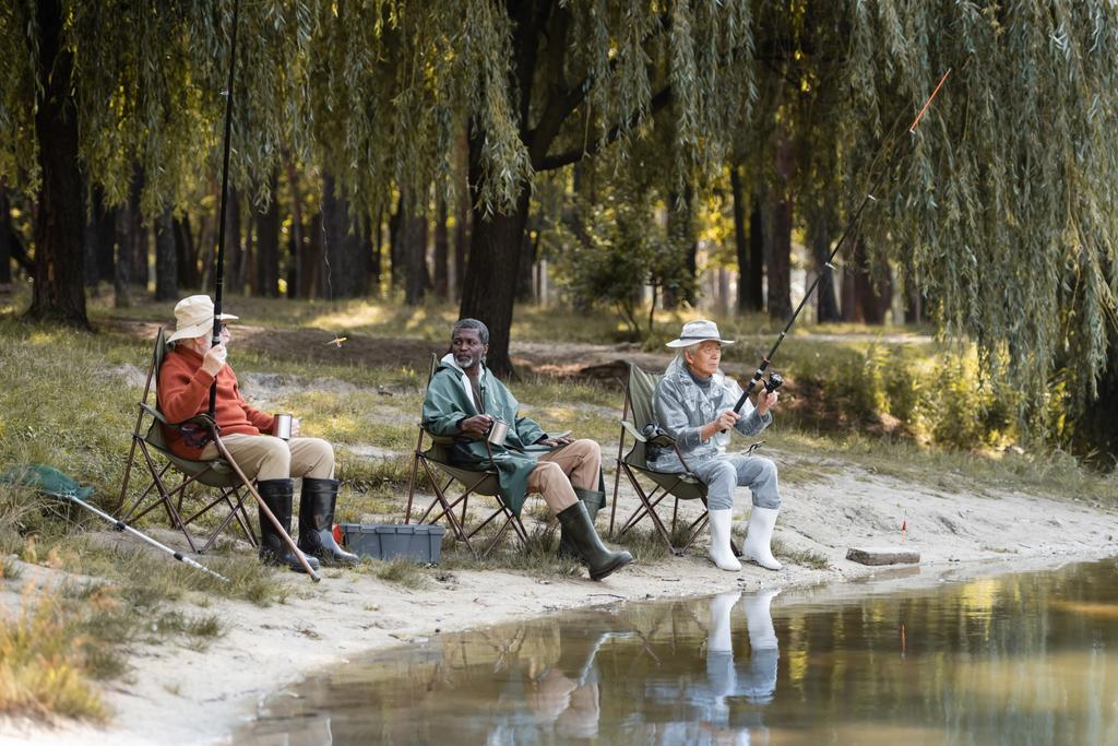 Πολυεθνικοί ανώτεροι άνδρες με καλάμια ψαρέματος που κρατούν θερμοφόρα κοντά στη λίμνη  - Φωτογραφία, εικόνα