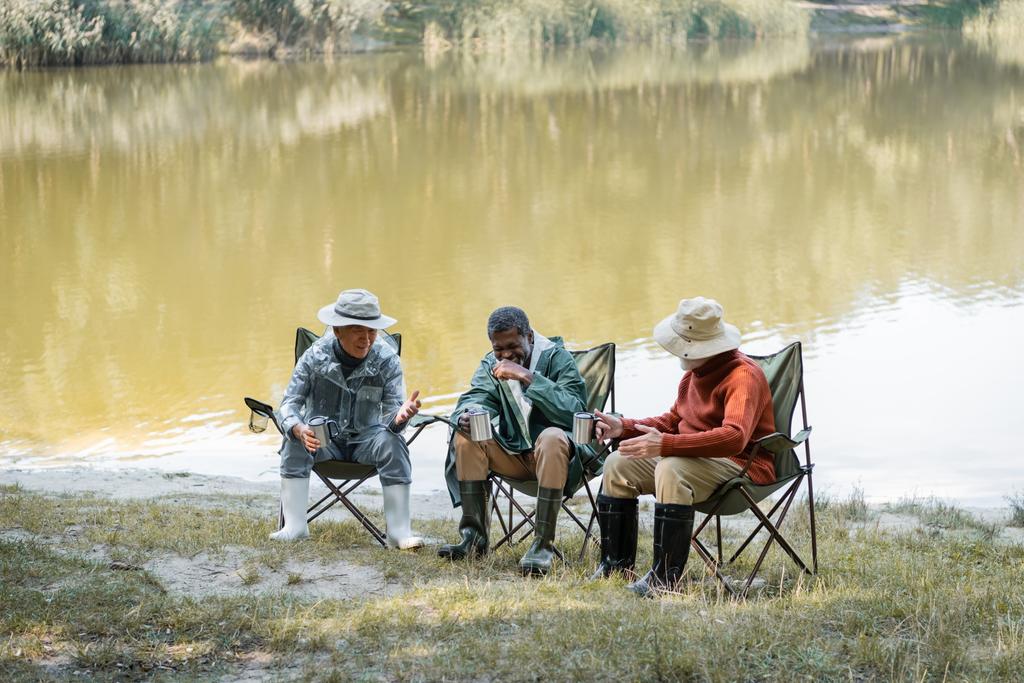 Positivo interracial senior hombres en traje de pesca celebración de termo tazas mientras pasar tiempo al aire libre  - Foto, imagen