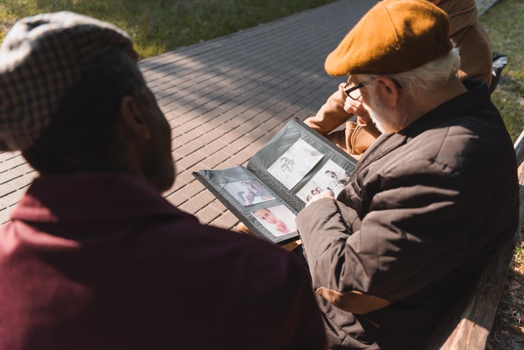 Elderly man holding photo album near blurred interracial friends on bench in park  - Photo, Image