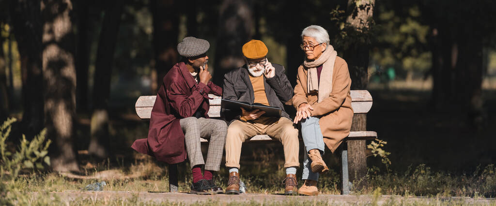 Senior man met fotoalbum praten op smartphone in de buurt van interraciale vrienden in het park, banner  - Foto, afbeelding