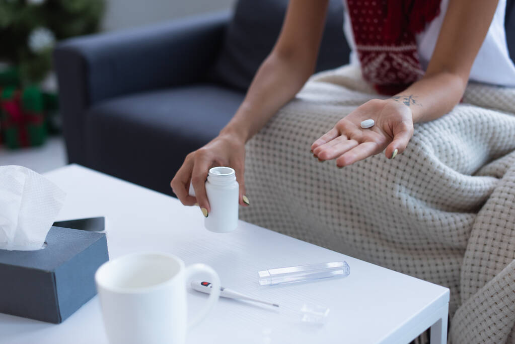 bijgesneden beeld van zieke vrouw met pil in de hand in de buurt van thermometer en een kopje warme drank op wazig tafel - Foto, afbeelding