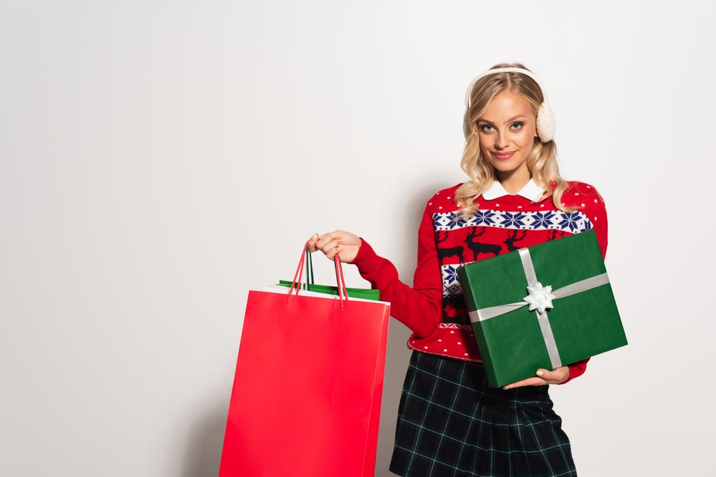 χαρούμενη γυναίκα με ωτοασπίδες και ζεστό πουλόβερ ποζάρουν με τσάντες για ψώνια και κουτί δώρου σε λευκό - Φωτογραφία, εικόνα