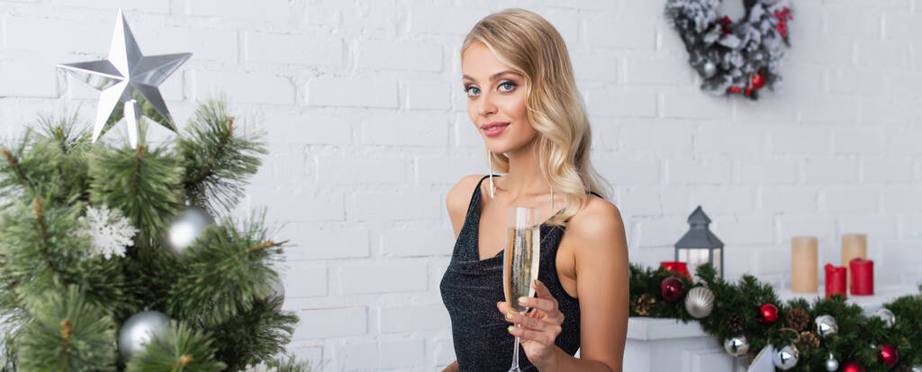 stylová blondýnka se sklenkou šampaňského při pohledu do kamery poblíž vánočního stromku, banner - Fotografie, Obrázek