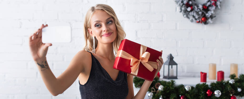 усміхнена жінка бере селфі з різдвяним подарунком у прикрашеній вітальні, банер
 - Фото, зображення