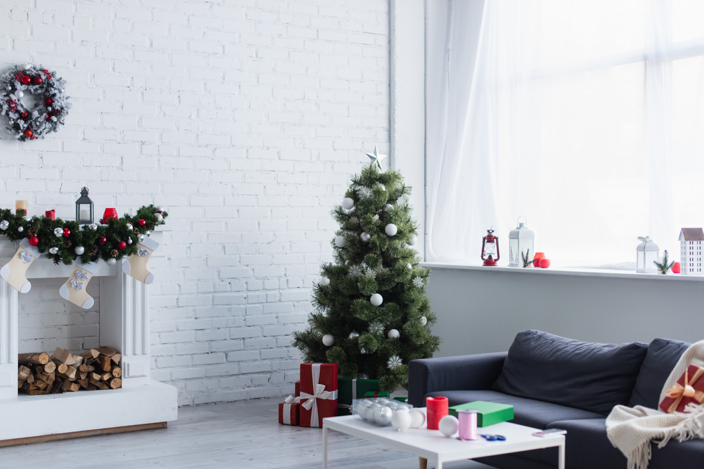 przestronny salon z choinką, ozdobiony kominkiem, sofą i stołem z kulkami świątecznymi i kokardką ozdobnej wstążki - Zdjęcie, obraz