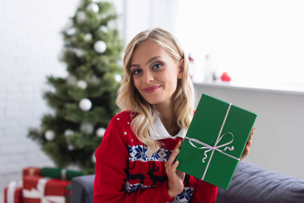 femme blonde gaie dans un pull chaud tenant boîte cadeau verte près de l'arbre de Noël sur fond flou - Photo, image