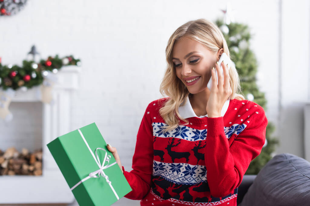 femme souriante en pull élégant avec ornement tenant cadeau de Noël tout en parlant sur smartphone - Photo, image