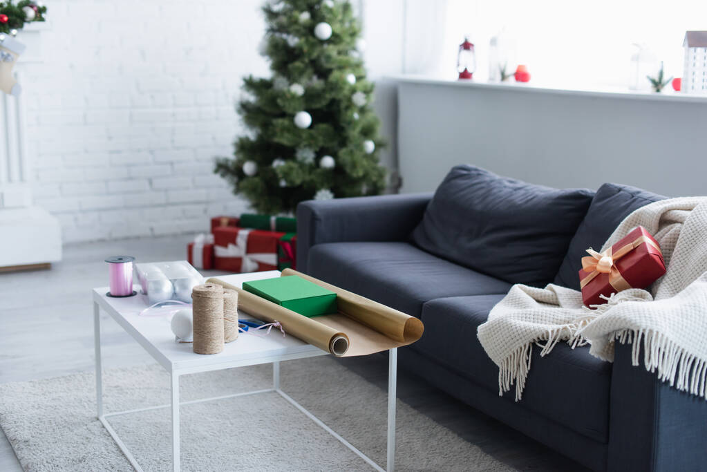 диван с теплым одеялом и подарочной коробкой возле стола с бечевкой, декоративной лентой и оберточной бумагой рядом с размытой елкой - Фото, изображение