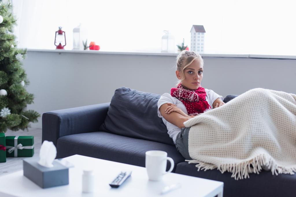 хвора жінка на дивані під теплою ковдрою біля ялинки на фоні
 - Фото, зображення