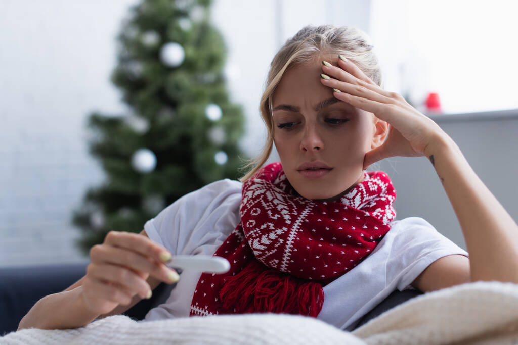 bezorgde vrouw in warme sjaal kijken naar thermometer in de buurt van kerstboom op wazig achtergrond - Foto, afbeelding
