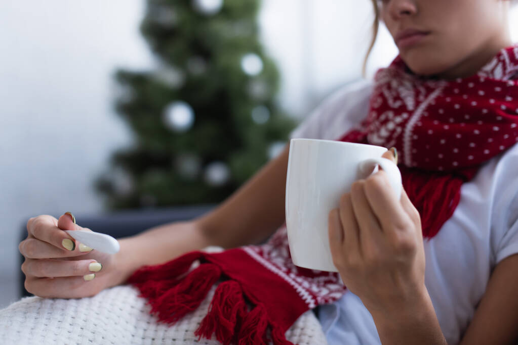 cropped view of sick woman with thermometer and cup of warm beverage near christmas tree on blurred background - Photo, Image