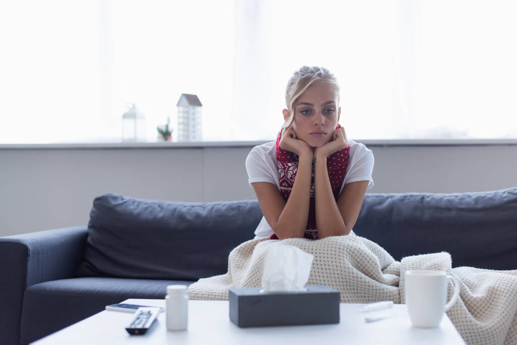 donna triste e malata in sciarpa calda seduta sul divano sotto coperta vicino a tovaglioli di carta e pillole contenitore - Foto, immagini