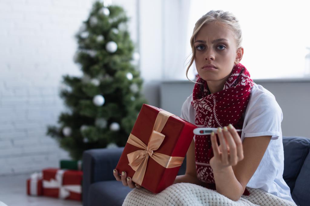 trieste vrouw met geschenkdoos en thermometer kijken naar camera in de buurt van kerstboom en presenteert op wazig achtergrond - Foto, afbeelding