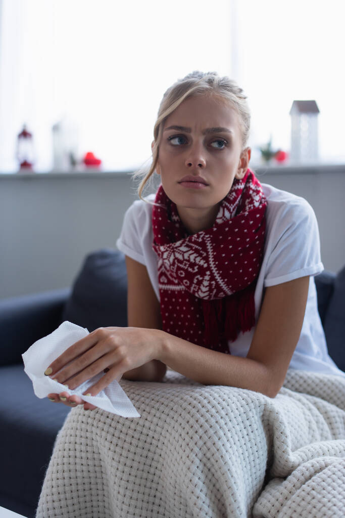 mujer enferma y pensativa en bufanda caliente sentado en el sofá con servilleta de papel  - Foto, Imagen