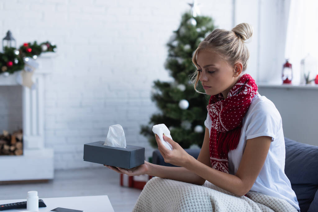 mujer enferma en bufanda caliente sosteniendo servilletas de papel cerca del árbol de Navidad sobre fondo borroso - Foto, imagen