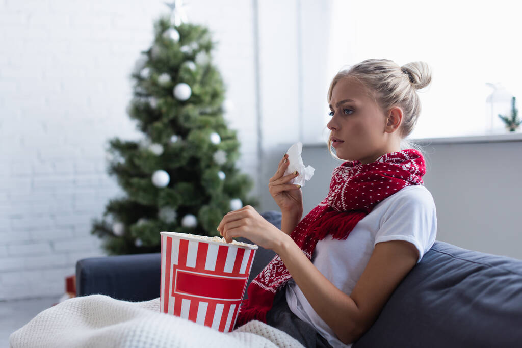 Kranke Frau mit Papierserviette und Eimer Popcorn vor dem Fernseher auf Sofa neben verschwommenem Weihnachtsbaum - Foto, Bild