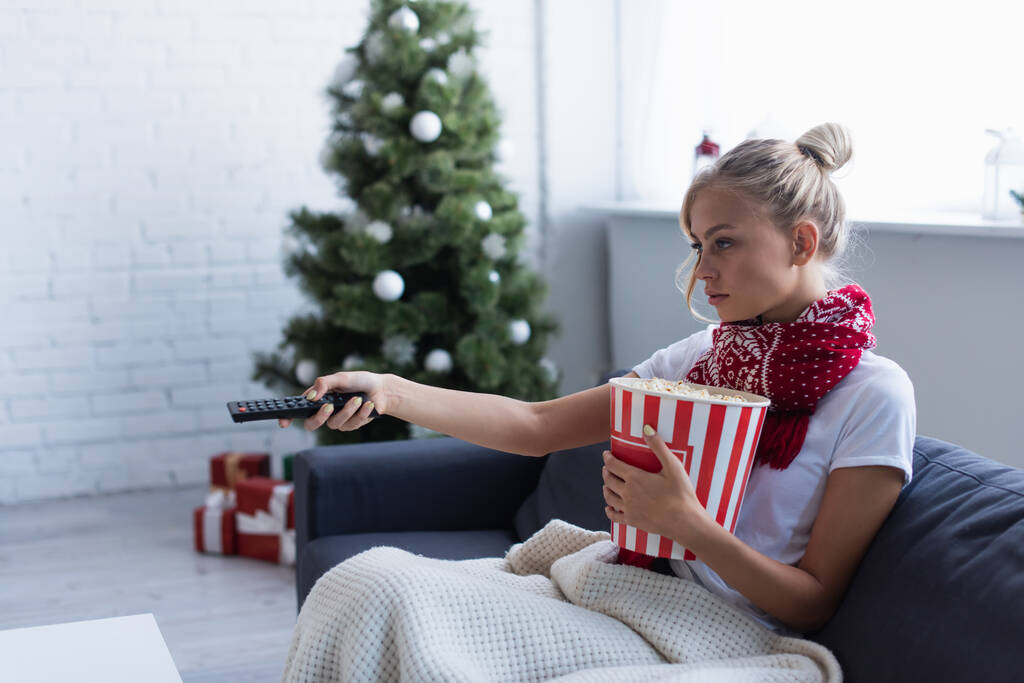 zieke vrouw met emmer van popcorn klikken kanalen terwijl het kijken van tv in de buurt wazig kerstboom - Foto, afbeelding
