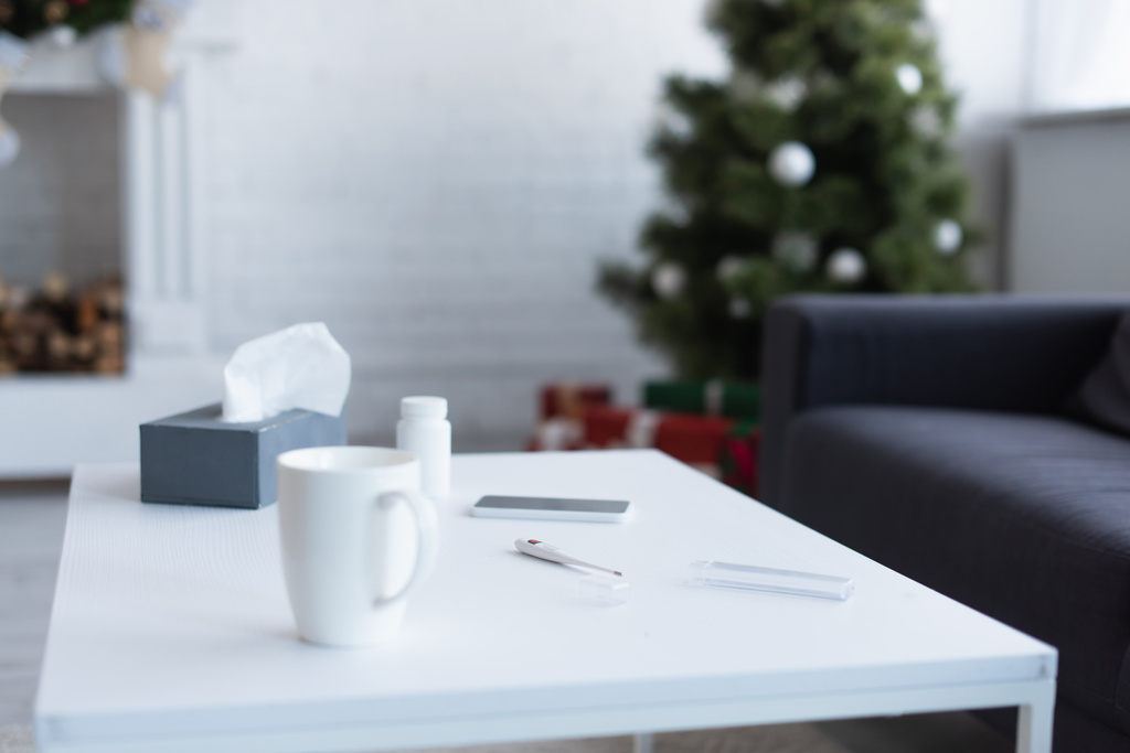 mesa com guardanapos de papel, recipiente de pílulas, termômetro eletrônico e smartphone perto da árvore de natal borrada - Foto, Imagem