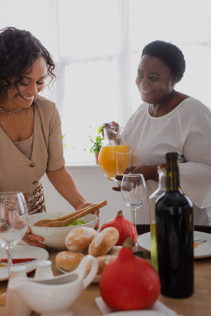 Mujer afroamericana sonriente con jugo de naranja mirando a su hija sirviendo la cena de Acción de Gracias  - Foto, Imagen