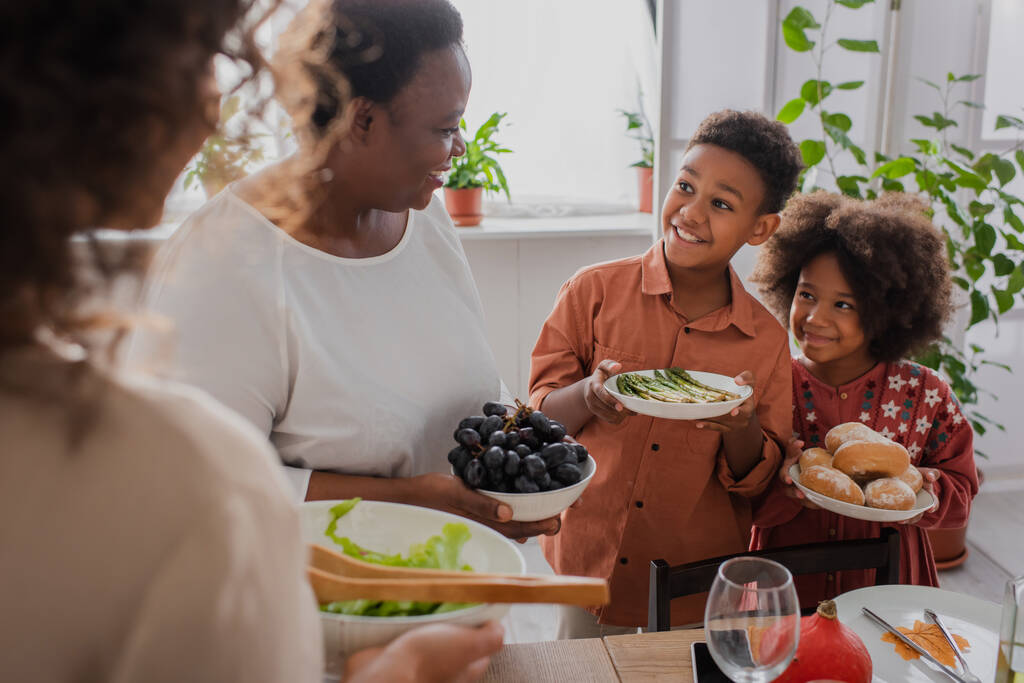 glimlachende Afrikaans-Amerikaanse kinderen houden eten in de buurt van oma en moeder tijdens Thanksgiving diner  - Foto, afbeelding
